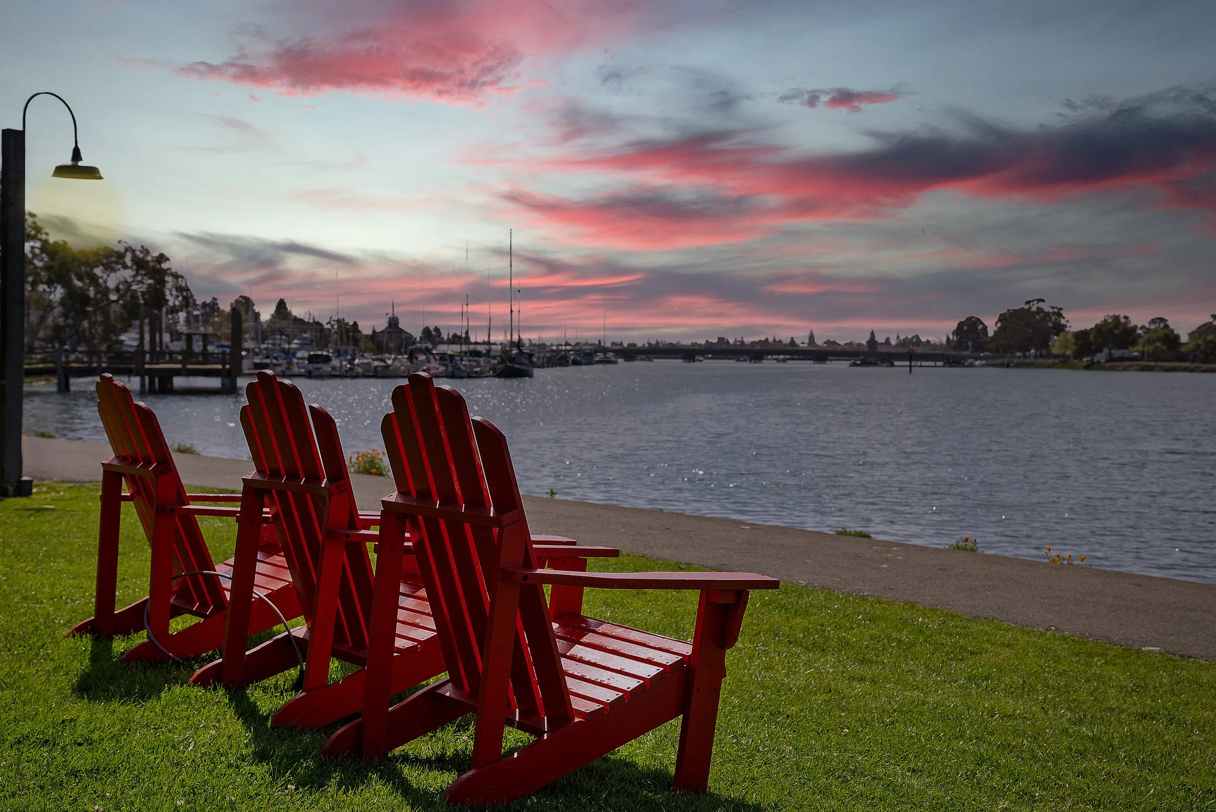 Three red Adirondack chairs on the lawn at Executive Inn & Suites, facing the Oakland estuary under a pink sunset